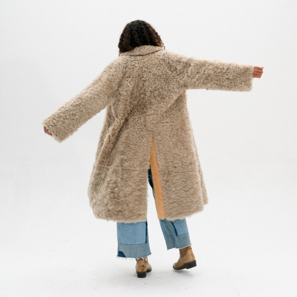 A person shown from behind wearing a long, shaggy beige teddy coat over light blue jeans and brown boots, standing against a plain white studio background with arms outstretched.