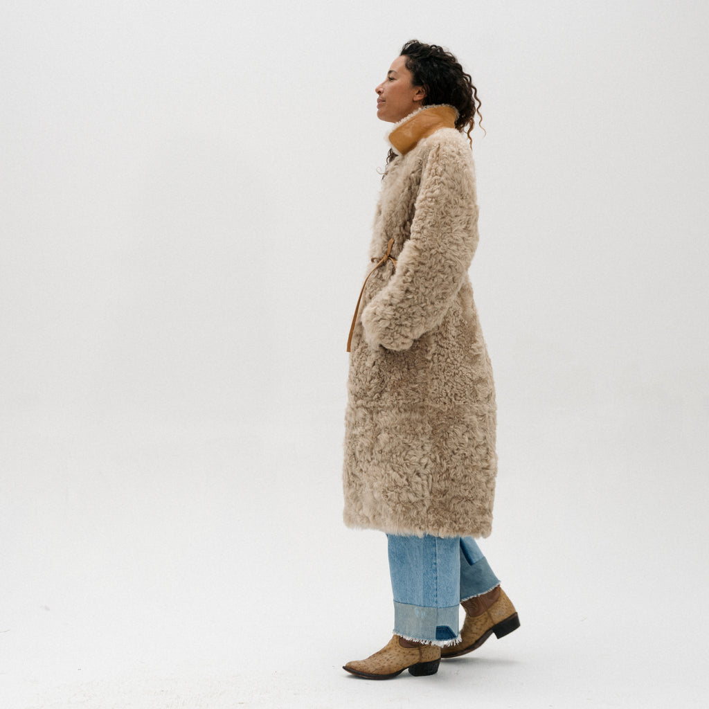 A woman in profile wearing a long, shaggy beige coat with a tan collar and belt, paired with light blue jeans and brown ankle boots, standing against a plain white studio background.
