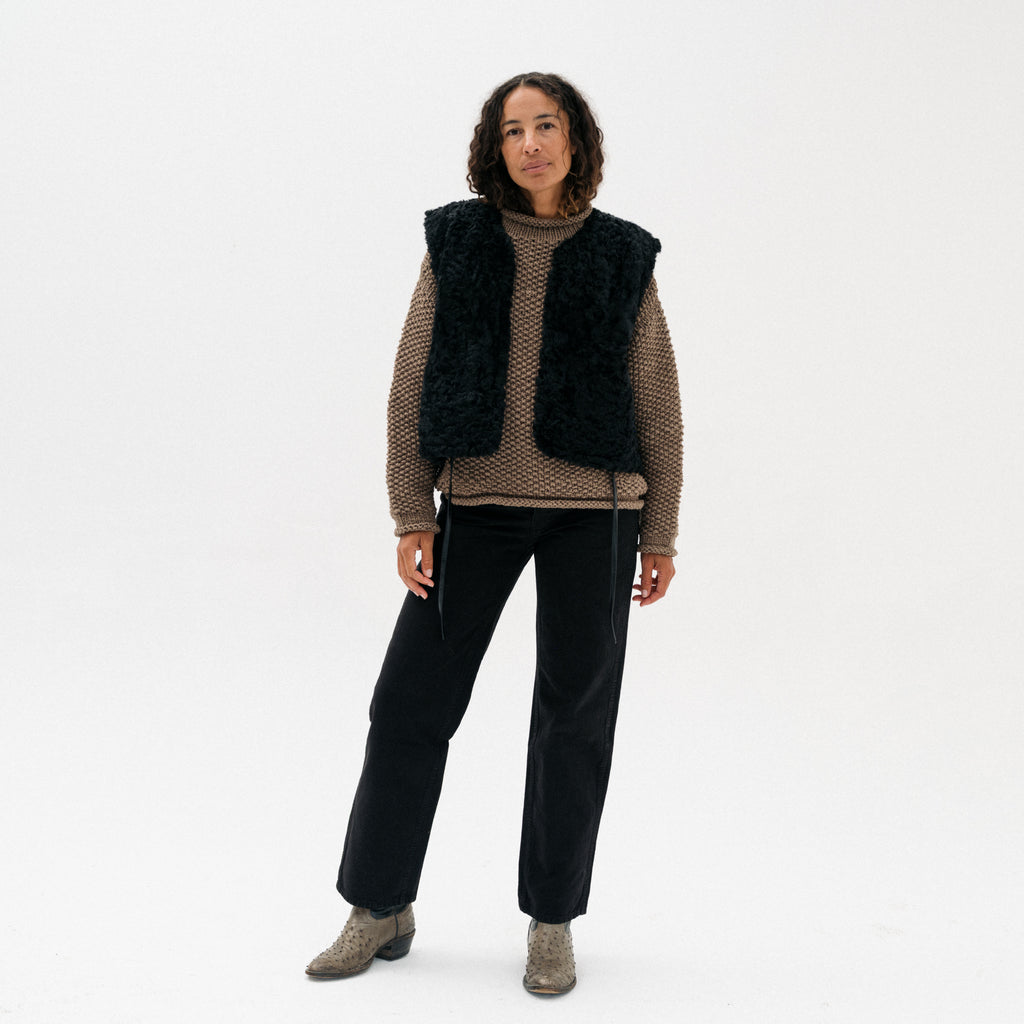 A model stands against a plain white studio background wearing a black fuzzy faux-fur vest over a brown textured knit sweater, black pants, and brown ankle boots.
