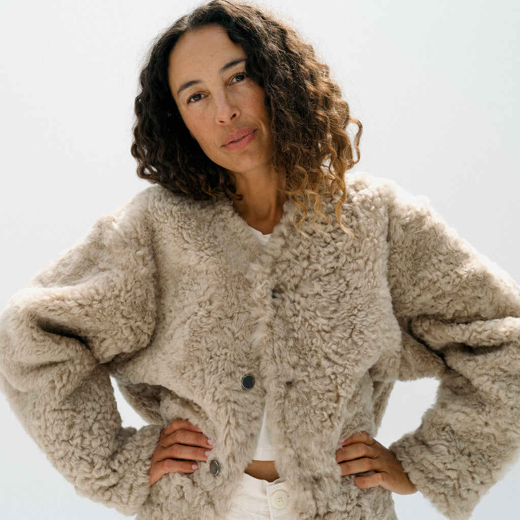 A woman with curly brown hair wearing a beige curly sherpa fleece cropped jacket over a white top, posing against a light gray studio background.