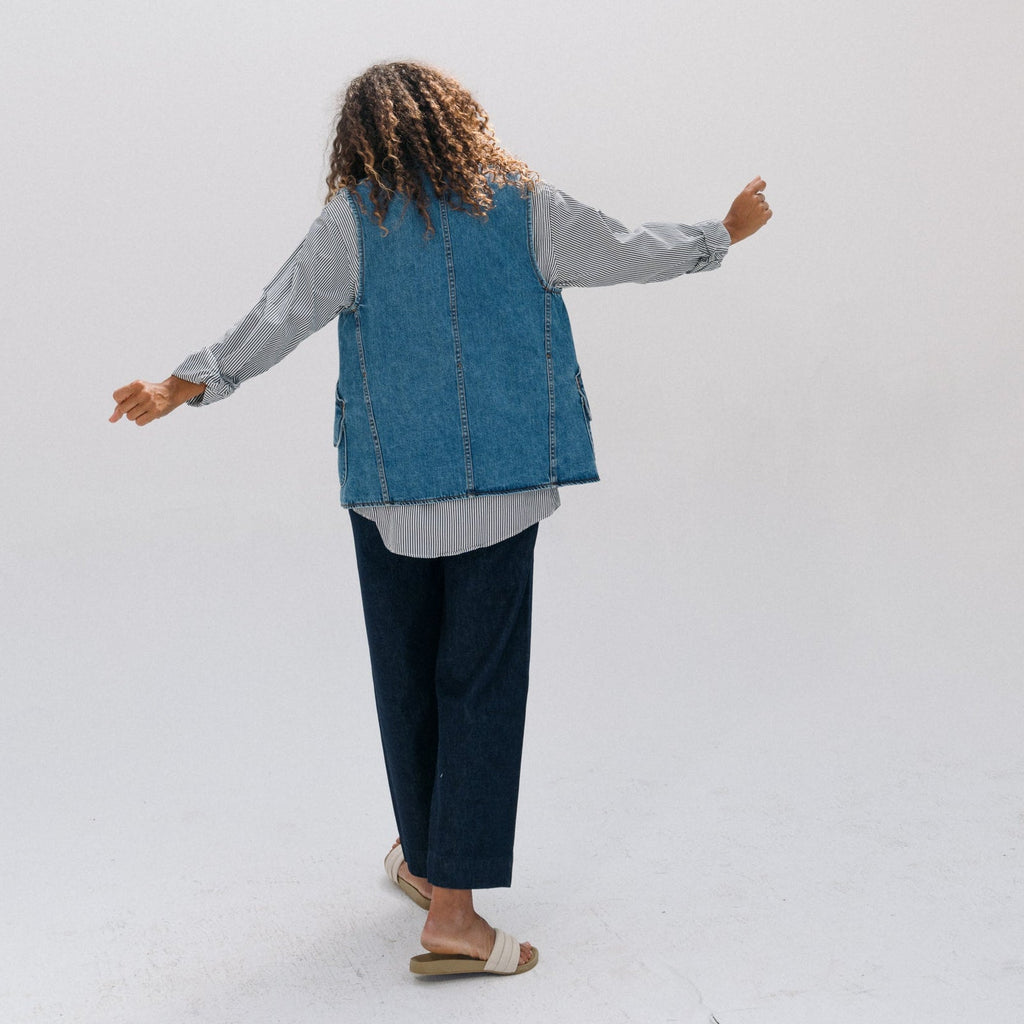 A person standing with arms outstretched, viewed from behind, wearing a blue denim vest over a light gray long-sleeve shirt and dark navy wide-leg pants, in a bright white studio setting.