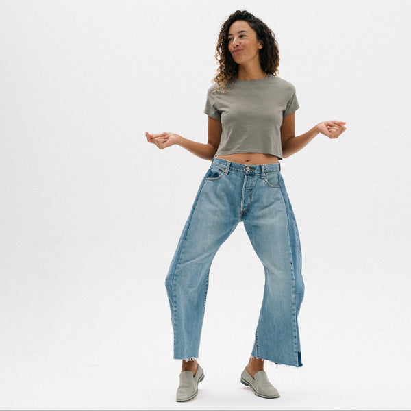 A woman modeling a pair of wide-leg, light-wash blue jeans with a raw hem, styled with a grey cropped t-shirt and grey slip-on shoes against a plain white studio background.