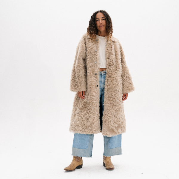 A woman modeling a long, shaggy beige teddy coat over a white crop top, light blue jeans, and tan boots, photographed against a seamless white studio background.