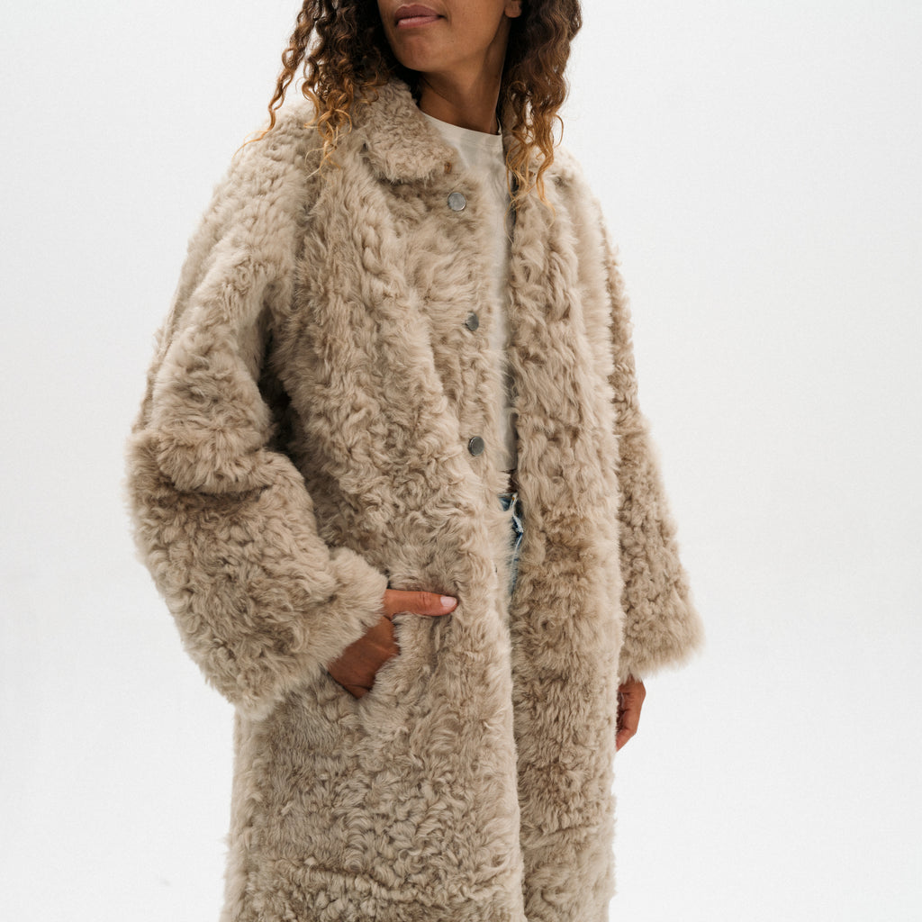 A woman wearing a long, shaggy beige teddy coat over a white top, photographed against a clean white studio background.