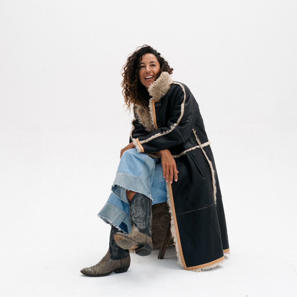 A woman sits on a stool wearing a long black shearling-lined coat with beige trim, a light blue denim skirt, and brown western boots against a plain white studio background.