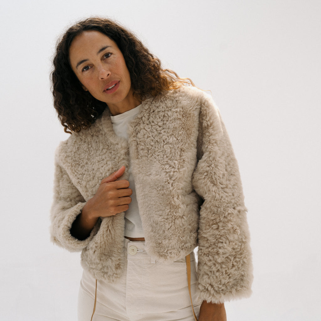 A woman modeling a cropped, textured beige sherpa jacket over a white top and white pants, photographed against a clean white studio background.