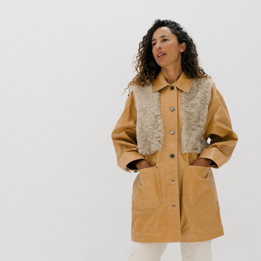 A woman modeling a tan jacket with a textured beige knit collar, paired with cream wide-leg pants and brown shoes, against a plain white studio background.
