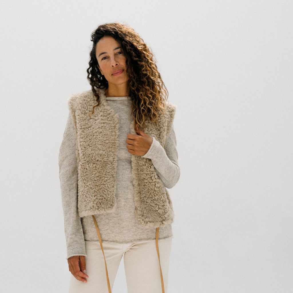 A woman modeling a beige textured faux-fur vest over a light gray knit sweater and cream drawstring pants, set against a seamless white studio background.