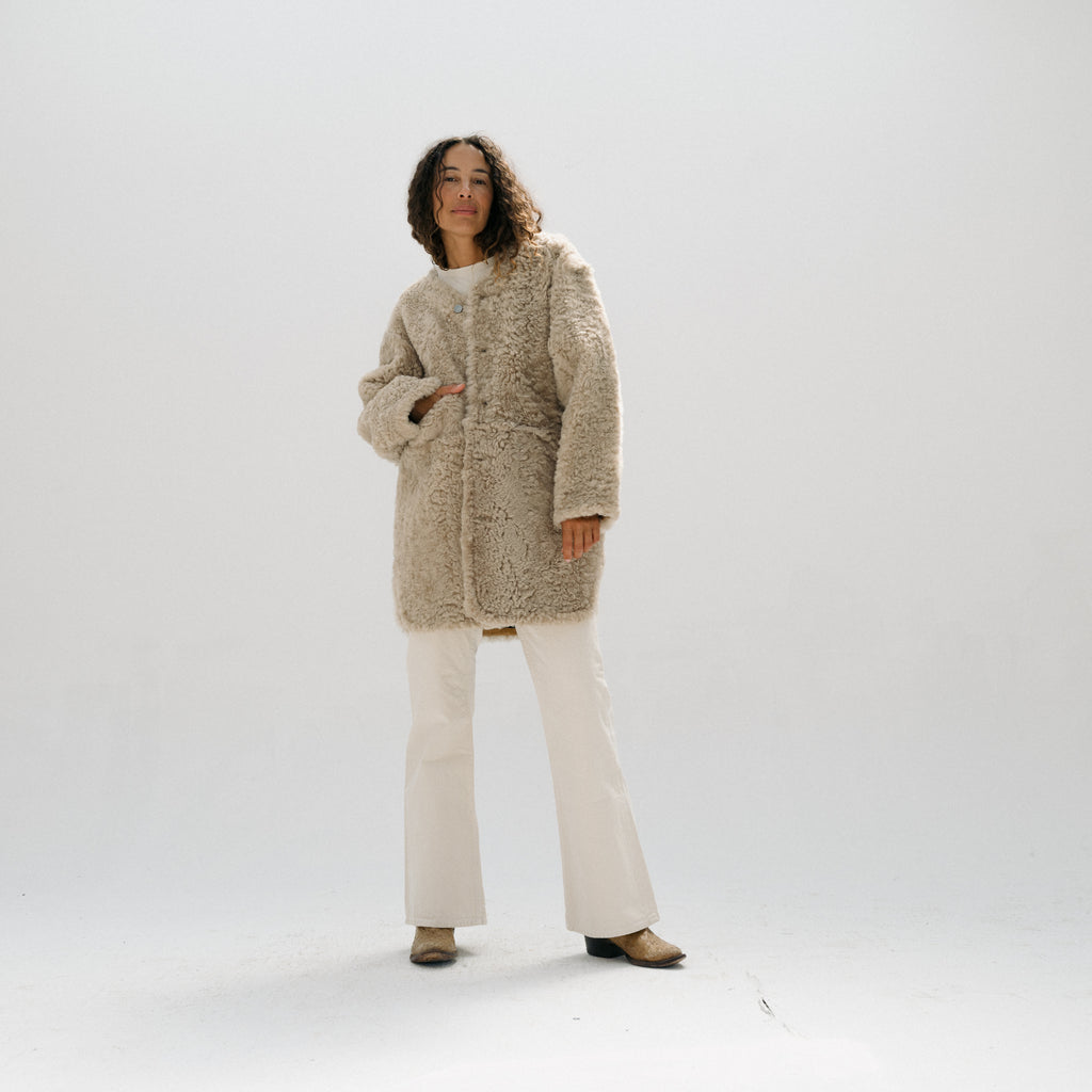 A model stands against a seamless white studio background wearing a textured, shaggy beige mid-length coat over a white top and white wide-leg pants.