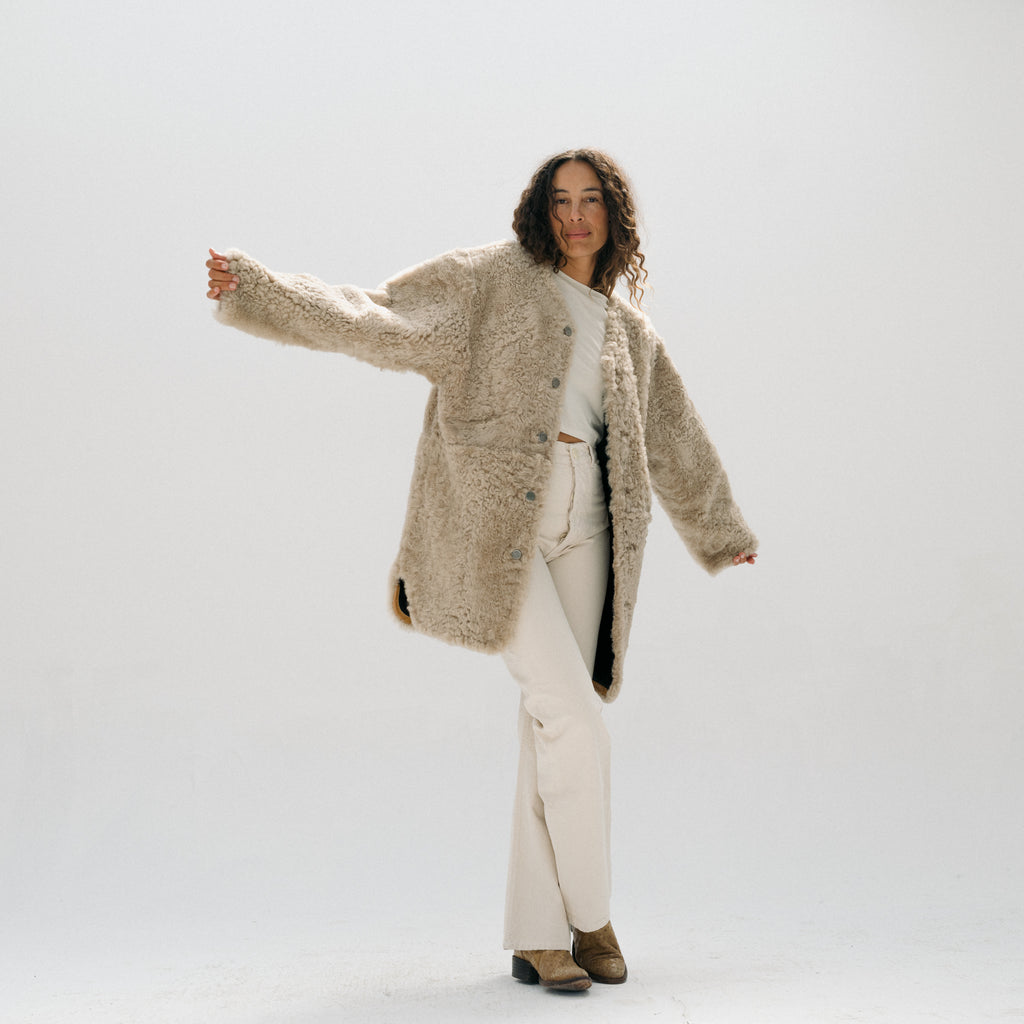 A woman modeling a beige shaggy faux-fur button-up coat over a white crop top and white wide-leg pants, standing against a seamless light gray studio background.