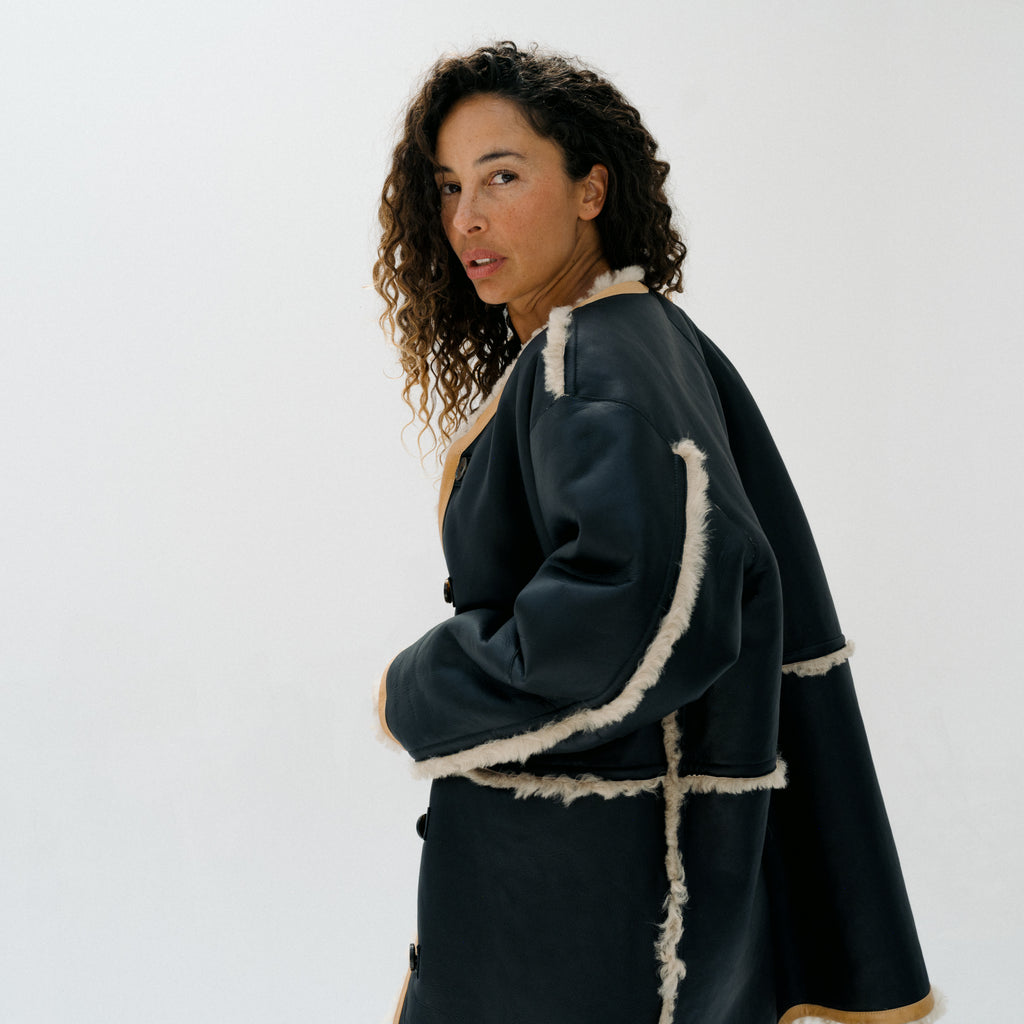 A woman wearing a dark navy blue coat with beige shearling trim, photographed against a plain white studio background.