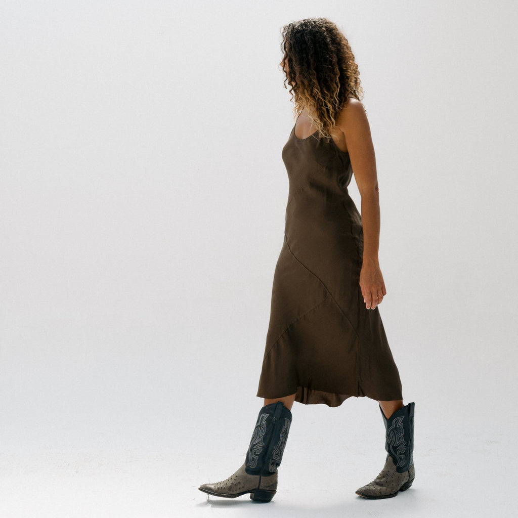A woman is shown from the back wearing a sleeveless brown midi dress and dark cowboy boots, standing against a seamless white studio background.